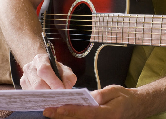 A man writing a song with a pen and paper. He is playing the acoustic guitar.